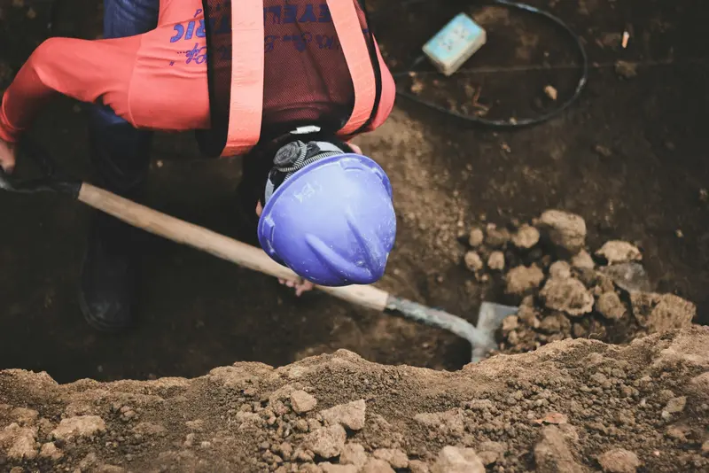A worker in a hard hat hand-digging soil at the base of an excavation